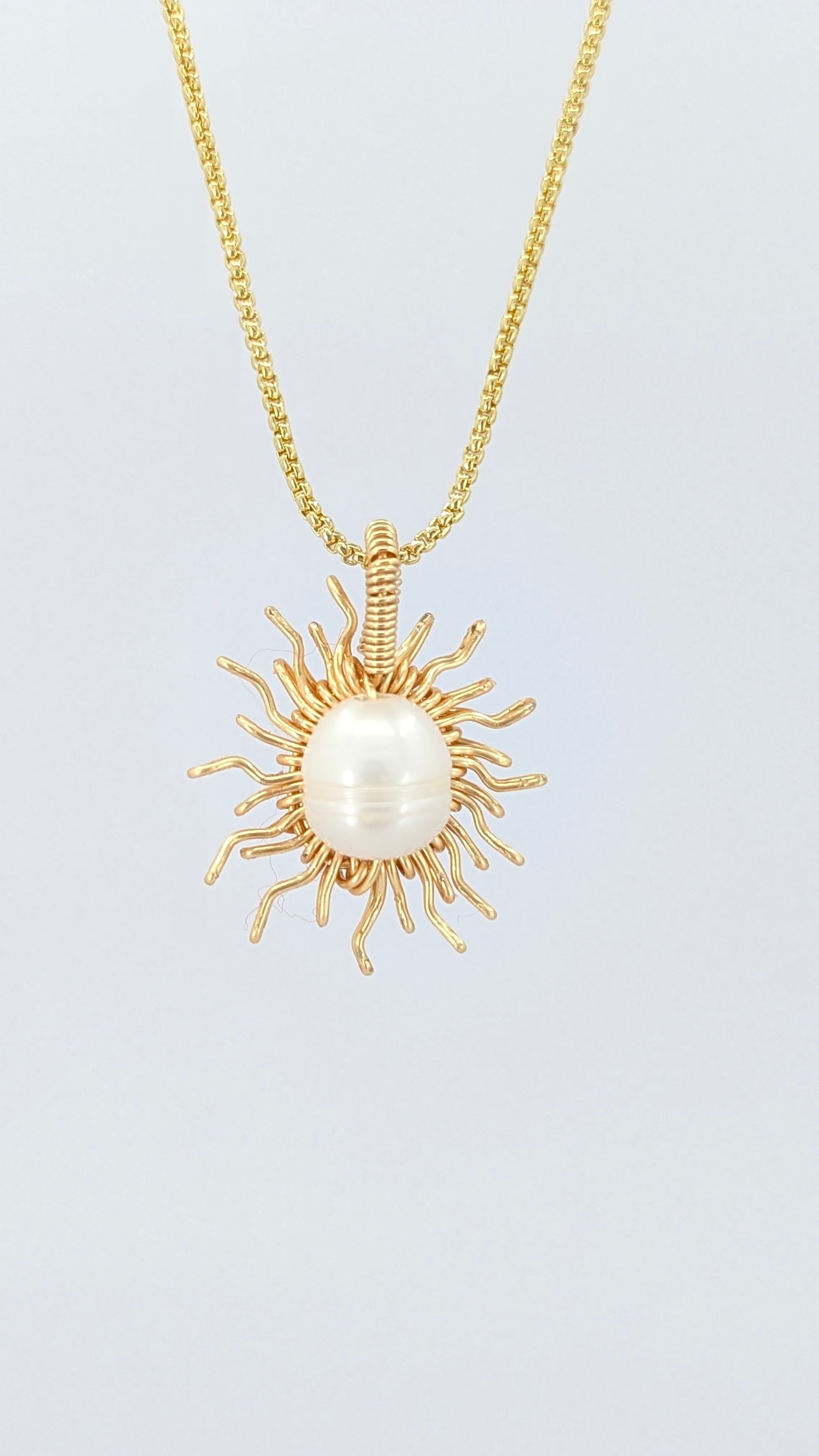 Front view of a gold necklace with a sun-shaped pendant featuring a pearl on a light gray background